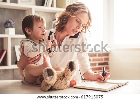 Beautiful business mom is talking on the mobile phone and taking notes while spending time with her cute baby boy at home Royalty-Free Stock Photo #617915075