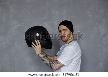 Latin handsome macho with beard and tattoo hands in white t-shirt keep black motorcycle helmet and look to the camera. Bikers concept for advertising motorbiker goods.