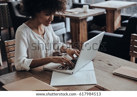 Developing new project. Beautiful young African woman using computer and smiling while sitting in cafe Royalty-Free Stock Photo #617984513