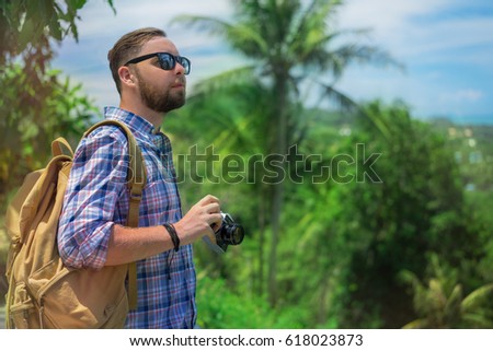 Young  male tourist with a backpack holding retro camera in his hands going to take next photo. Beautiful exotic landscape with the palm trees at the background. Mock up, copy space for your text.