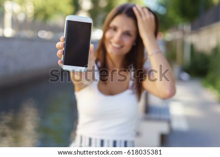 Young woman holding out her blank smartphone with the screen towards the camera as she stands alongside a river or canal in summer clothes, focus to the phone