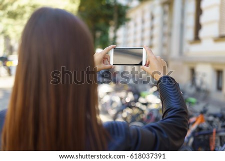 Young woman taking a photo on her mobile phone focusing on an old historic building with bicycles parked in front of it, over the shoulder view