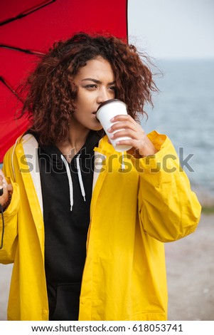Picture of concentrated african curly young lady wearing yellow coat holding umbrella walking outdoors. Looking aside and drinking coffee.