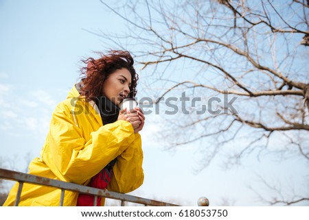 Picture of amazing african curly young lady wearing yellow coat walking outdoors. Looking aside and drinking coffee.