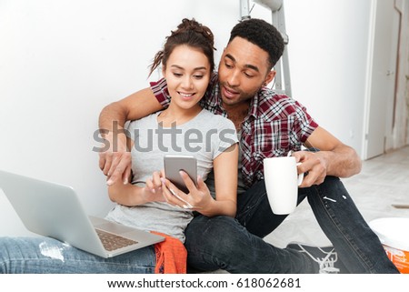 Picture of cheerful young loving couple sitting on floor in new flat makes repair and using laptop computer drinking tea. Looking aside while chatting by phone.