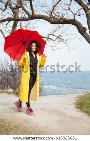 Picture of smiling african curly young lady wearing yellow coat holding umbrella walking outdoors. Looking at camera.