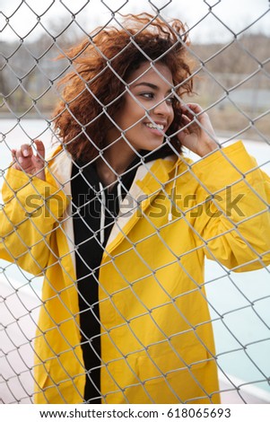 Picture of happy african curly young lady wearing yellow coat standing near fence outdoors. Looking aside.