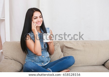 Beautiful woman sitting on the couch while holding a cup of hot tea and speaking by phone in the apartment