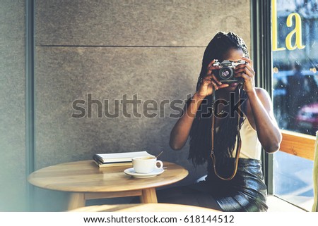 Afro american female traveller making photo of cafe interior on vintage camera sitting at wooden table with diary for interesting places and cup of tasty coffee.Copy space for your advertising area