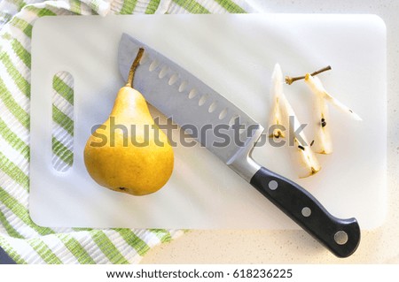A ripe delicious yellow pear on a white plastic chopping board together with a chef's knife and leftovers and stubs of another pear