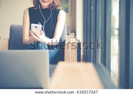 Charming beautiful woman is listening music by her smartphone and feeling chill at coffee shop cafe. Close-up of her phone.