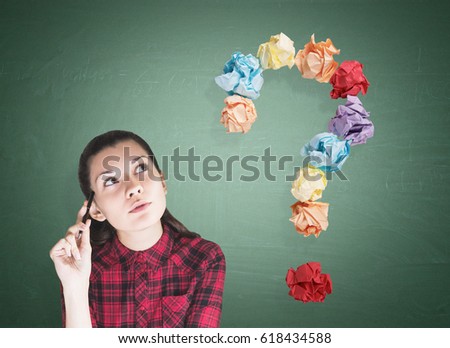 Portrait of a geeky young woman wearing a red checkered shirt and sitting with a pen near a green chalkboard. There is a large question mark made of colorful paper balls to the left of her.