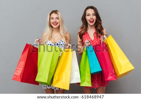 Picture of happy young two ladies friends with bright makeup lips standing over grey wall and posing with shopping bags. Looking at camera.