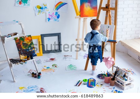 Cute, serious and focused, white three years old boy in blue shirt and jeans apron drawing on canvas standing on the easel. Concept of early childhood education, talent, happy family or parenting Royalty-Free Stock Photo #618489863