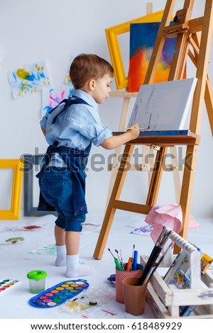 Cute, serious and focused, white three years old boy in blue shirt and jeans apron drawing on canvas standing on the easel. Concept of early childhood education, talent, happy family or parenting Royalty-Free Stock Photo #618489929