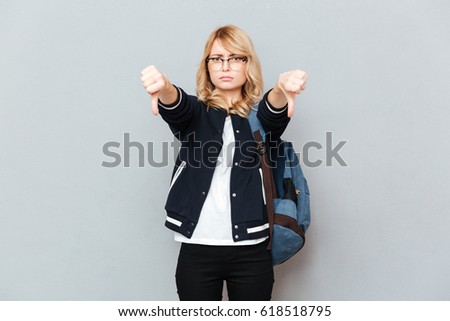Photo of sad young lady student wearing glasses with backpack posing over grey wall. Looking at camera showing thumbs down.