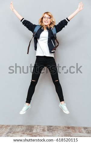 Photo of happy young lady student wearing glasses with backpack jumping over grey wall. Looking at camera.