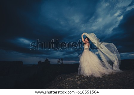 Dreamy and mysterious picture of a lost bride on a mountain rock, with her dress floating into the wind and on the background a dark ancient castle