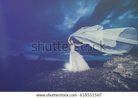 Dreamy and mysterious picture of a lost bride on a mountain rock, with her dress floating into the wind and on the background a dark ancient castle