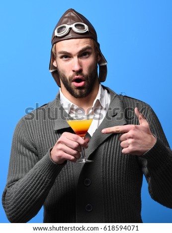 handsome bearded aviator man with beard and surprised face holding glass of alcoholic cocktail in gray knitted sweater with hat and glasses on blue studio background