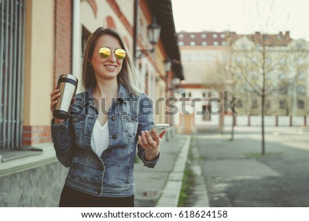 Happy young hipster woman walking on the street, using her smartphone and drinking coffee