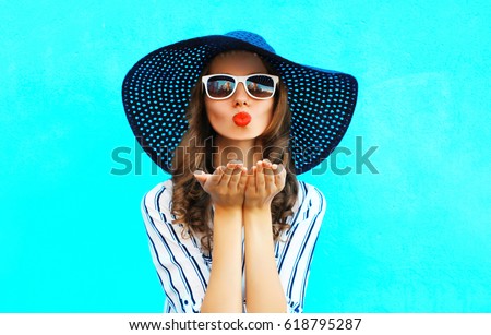 Portrait of beautiful young woman blowing her lips sending air kiss wearing sunglasses, summer straw hat on blue background