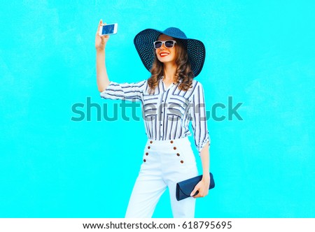 Fashion young smiling woman is taking a picture on a smartphone wearing a straw hat, white pants with a handbag clutch over colorful blue background