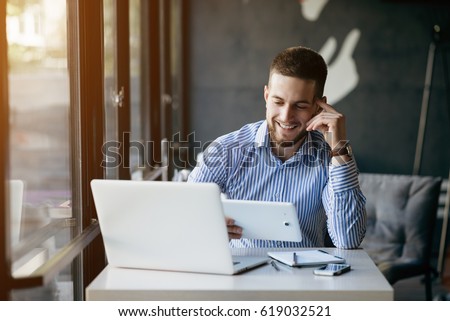 Young handsome man sitting in office with cup of coffee and working on project connected with modern cyber technologies. Businessman with notebook trying to keep deadline in digital marketing sphere.