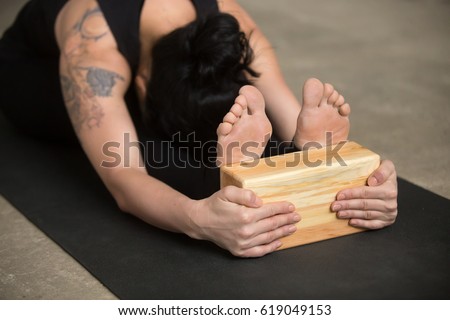 Young sporty yogi woman practicing yoga concept, sitting in paschimottanasana exercise using block, doing seated forward bend pose, working out, close up of feet and hands, tattoo on shoulder 