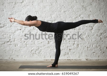 Young yogi attractive woman practicing yoga concept, standing in Warrior three exercise, Virabhadrasana III pose, working out, wearing sportswear, black pants, full length, white loft background 