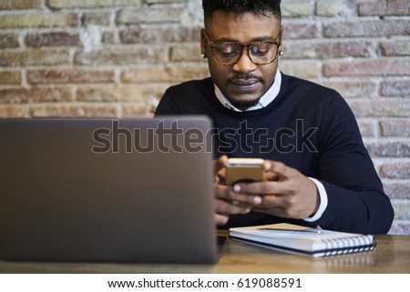 Cropped image of afro American hipster guy in eyeglasses doing shopping on internet websites on mobile phone using free internet connection. Hipster man downloading new mobile application on cellular
