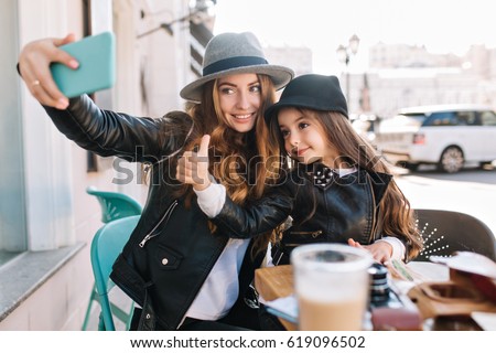  Stylish family sitting in a city cafe look in the phone and take selfies and smile on the sunny city background. Little girl show finger to the top looking at camera. True emotions,  good mood.