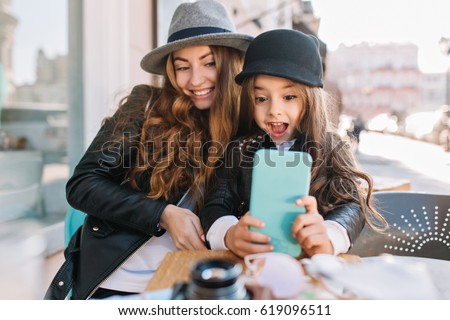 Pretty young mother and her cute daughter having fun and  take selfies. Little girl surprised looking in phone and smile on the sunny city background. Stylish family, true emotion, good moon.