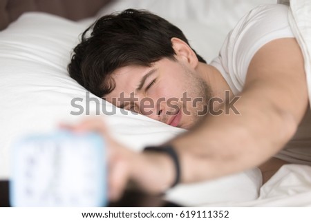 Sleepy guy waking up early after hearing alarm clock signal on monday morning, with eyes still closed reaching button on the clock to turn it off, feeling tired and does not want to get up  Royalty-Free Stock Photo #619111352