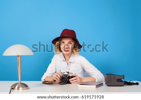 Happy girl photographer sitting at the table.Young beautiful girl in white dress holding camera and sitting at white table isolated on blue background.Hipster girl dressed in casual wear,red hat,curly