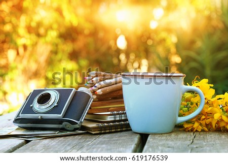 Image of notebooks, vintage photo camera, colorful pencils next to field flowers on wooden table outdoors at afternoon. selective focus