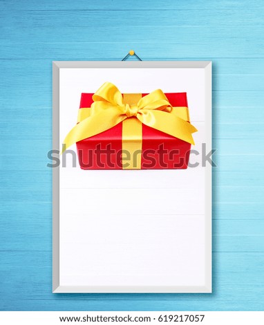 Gift box with yellow bow. Present wrapped with ribbon. Christmas or birthday red package. On white wooden table. Photo frame on wooden rustic wall.