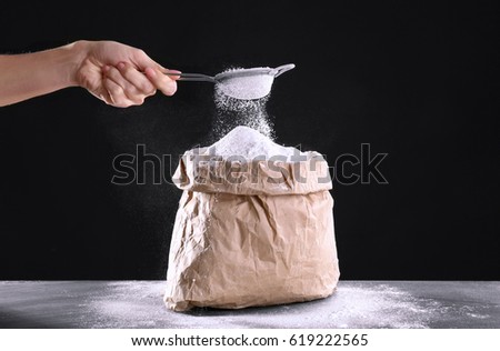 Bag of flour and female hand with sieve on dark background