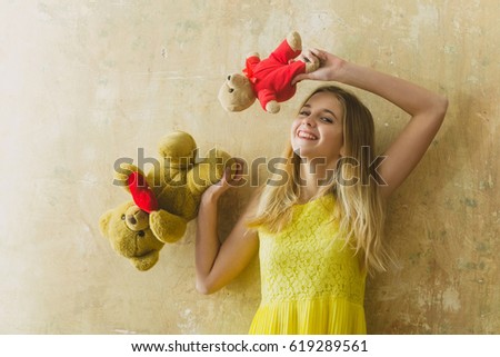 Pretty girl or cute smiling woman with blond hair and adorable happy face in yellow dress with teddy bear toy for valentines day on textured wall background, copy space