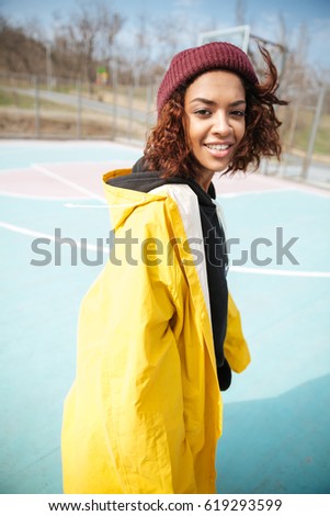 Picture of smiling african curly young woman wearing yellow coat walking outdoors. Looking at camera.