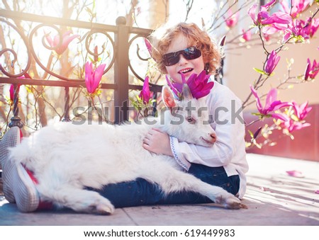 smiling boy with curly hair and white goat, blossom magnolia flowers on background, goatling and boy, little friends, sunny day, spring morning, best friends on spring background.
