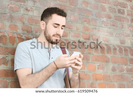 Portrait of young latin man typing on his phone against brick wall. Outdoors.