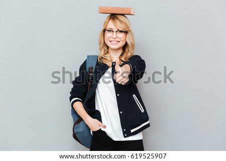 Happy female nerd in funny eyeglasses with book on head showing thumb up