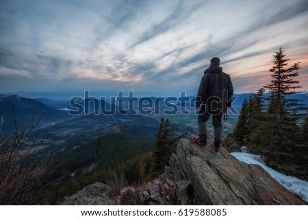 Man watching a beautiful cloudy sunset over a rocky cliff. Picture taken on top of Elk Mountain near Chilliwack, British Columbia, Canada.