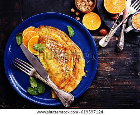 French pancakes - Crepe Suzette with orange syrup and hazelnuts on black wooden board. top view, copy space. Royalty-Free Stock Photo #619597124