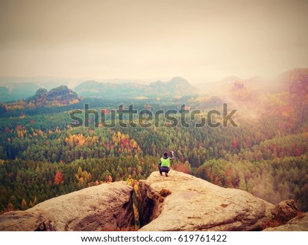 Hobby photographer takes picture of spring nature park from sharp rocks. Hiker in green jacket stay with camera on tripod on stony peak increased from foggy valley. 