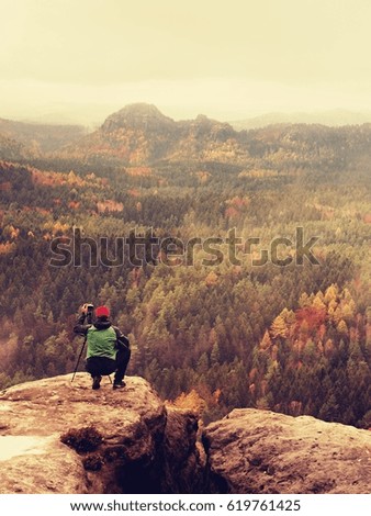 Hobby photographer takes picture of spring nature park from sharp rocks. Hiker in green jacket stay with camera on tripod on stony peak increased from foggy valley. 