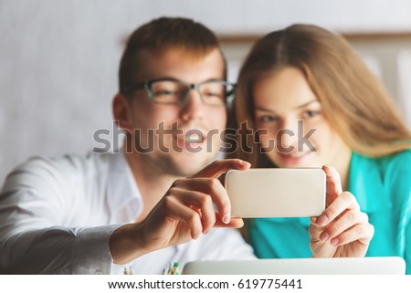 Blurry young man and woman at workplace taking selfie with smartphone. Social media concept