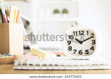 Close up of white clock placed on office desk with spiral notepad stickers, pencils, blurry laptop in the background and other items