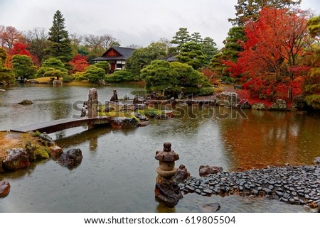 Autumn scenery of a beautiful Japanese garden in Katsura Imperial Villa ( Royal Park ) in Kyoto, Japan, with view of fiery maple trees by the lake and a stone bridge across the pond on a drizzling day